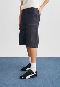 Oseba, ki nosi temno modre kavbojke do kolen, bele nogavice in črne Puma superge, stoji na modrem preprogi.