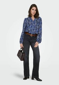 Femme portant un chemisier à motifs bleus et blancs, un jean taille haute foncé, des chaussures à talons noires, et portant un sac à main foncé.