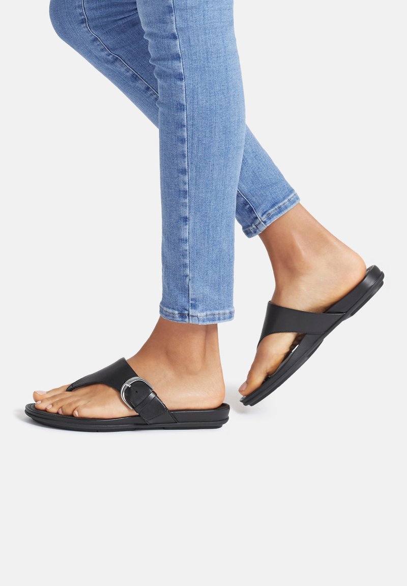 Black leather sandals with a buckle, featuring a toe post design. Flat sole, smooth finish, paired with light blue skinny jeans.