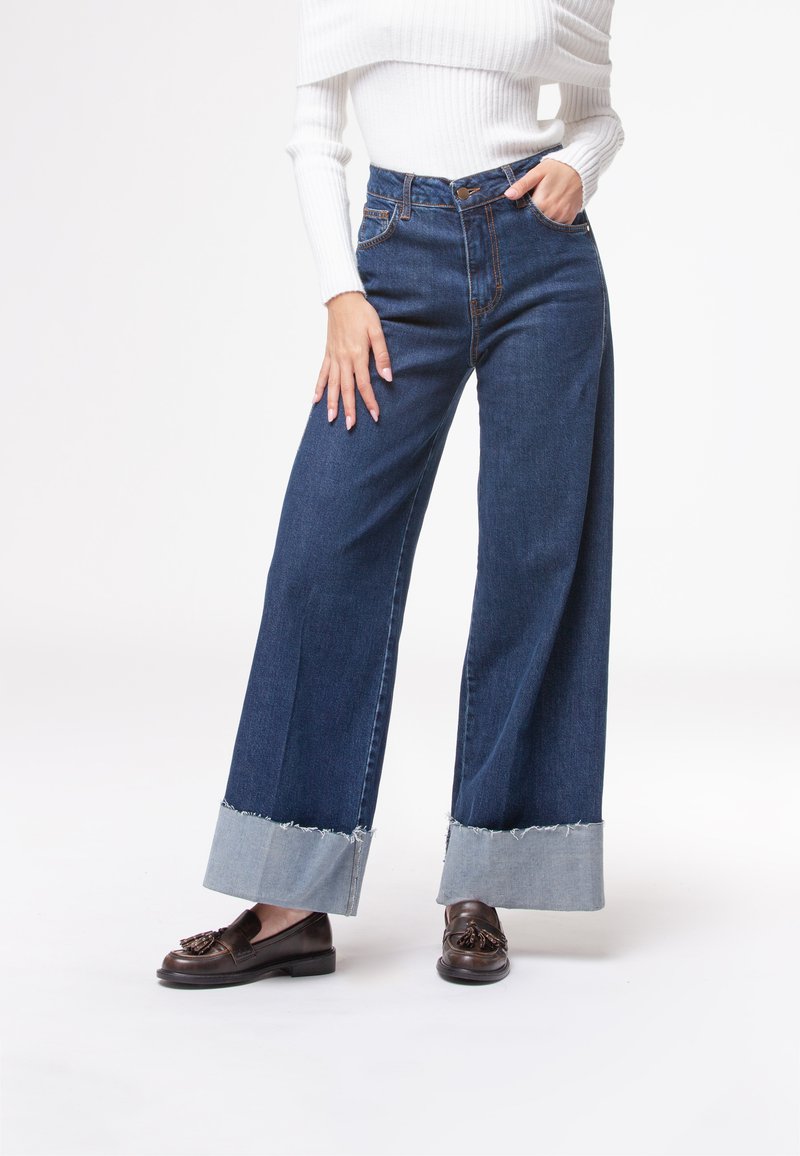 Donna che indossa jeans a gamba larga di colore blu scuro con risvolti chiari, un maglione bianco a coste off-shoulder e mocassini marrone scuro con frange.
