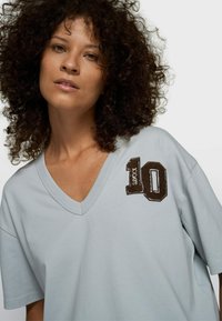 Camiseta de algodón azul claro con cuello en V. Presenta un número "10" bordado en marrón texturizado con una fuente deportiva en el área del pecho izquierdo.