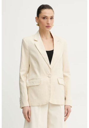 Donna con capelli tirati indietro che indossa orecchini a cerchio dorati, un blazer crema sopra una maglietta nera e pantaloni crema, in piedi davanti a uno sfondo bianco.