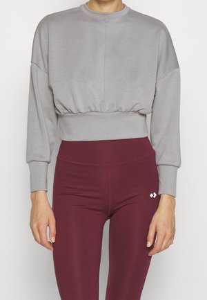 Sudadera gris recortada con cuello redondo, hombros caídos y cintura elástica, combinada con leggings marrón burdeos que presentan un pequeño logo blanco.