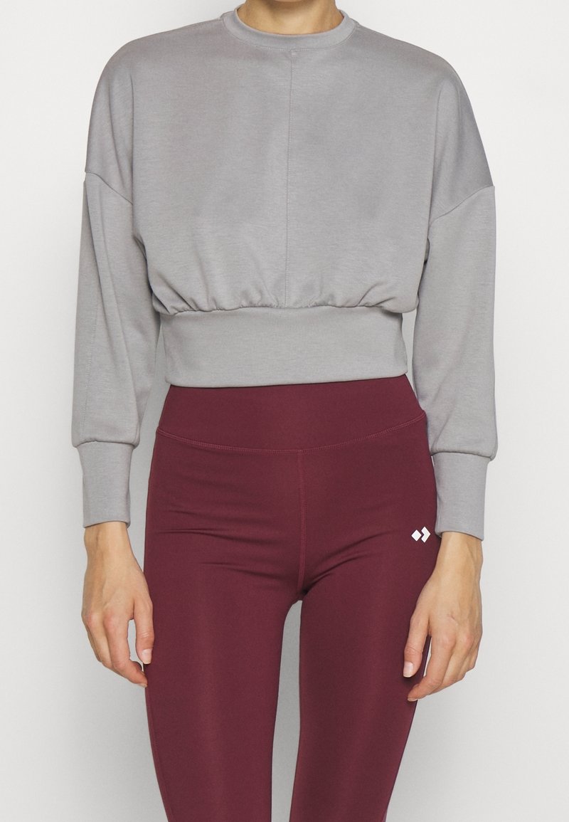 Felpa grigia corta con colletto rotondo, spalle basse e vita elasticizzata, abbinata a leggings bordeaux con un piccolo logo bianco.