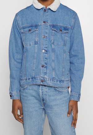 Mies pukeutuneena vaaleansiniseen farkkutakkiin, jossa on valkoinen sherpa-kaulus, ja vastaaviin vaaleansinisiin farkkuihin, seisoo valkoista taustaa vasten.