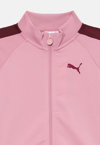 Roze sportjack met een hoge kraag, met een kastanjebruine panel op de schouders en een klein geborduurd logo op de linkerborst.