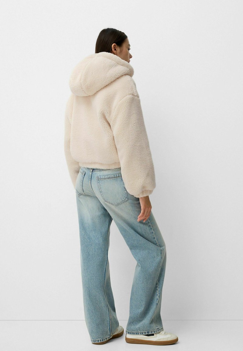 Un hoodie en polaire beige clair avec une capuche arrondie, une texture douce et des poignets élastiques, associé à un jean large bleu clair et des baskets crème.