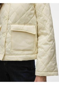 Chaqueta de crema acolchada con cuatro botones, un bolsillo en el pecho y mangas anchas. La tela parece suave con un brillo sutil y un patrón de diamantes.