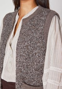 Gilet lavorato a maglia con un corpo marrone texturizzato e bordi a costine, caratterizzato da un colletto a scialle e tasche frontali, indossato sopra una blusa a righe.