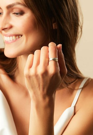 Lächelnde Frau mit braunen Haaren, die ein weißes Oberteil trägt, zeigt einen silbernen, gedrehten Ring an ihrer erhobenen Hand in der Nähe ihres Gesichts.