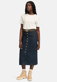 Kvinde med fletninger iført en hvid t-shirt, en knaplukket mørk denim midi-nederdel med lommer, brun bælte og beige støvler med sokker.