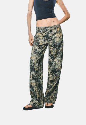 Persona delgada con top corto negro, pantalones sueltos con patrón tie-dye y chanclas negras, de pie con una mano detrás de la espalda.