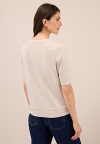 Kvinde med langt brunt hår iført en beige kortærmet skjorte og mørkeblå jeans, vendt sidelæns mod en ensfarvet baggrund.