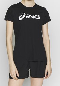Person, der ein schwarzes ASICS T-Shirt und passende schwarze Shorts trägt, steht mit entspannten Armen an den Seiten vor einem schlichten Hintergrund.