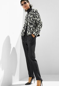 Vrouw in een zwart-witte patroonjas, zwarte coltrui, zwarte pantalon en zwarte hoge hakken, staande tegen een witte muur.