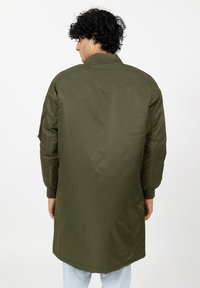 Man met krullend donker haar in een lange olijfgroene bomberjack, staand met zijn rug naar de camera tegen een witte achtergrond.