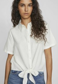 Camicia bianca a maniche corte in lino, con colletto e dettaglio annodato sul davanti. Abbinata a jeans in denim blu.