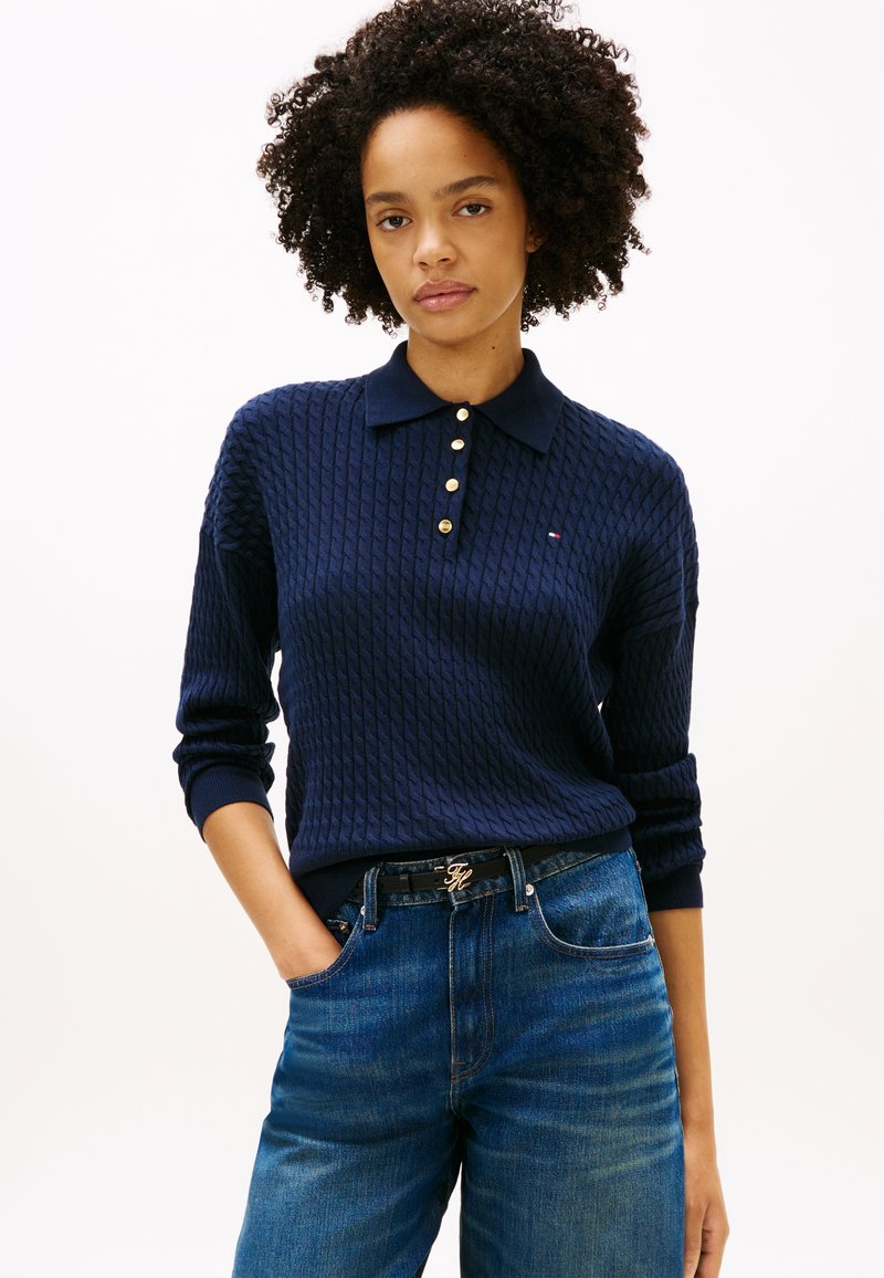 Femme aux cheveux bouclés portant un polo bleu marine texturé à manches longues et un jean bleu, posant avec une main dans la poche sur fond blanc.