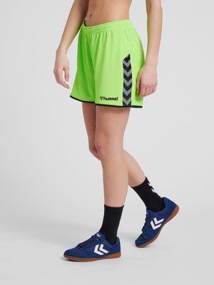 Person iført lysegrønne Hummel sportsshorts med sorte chevron-striber, sorte sokker og blå sneakers står på gråt gulv.