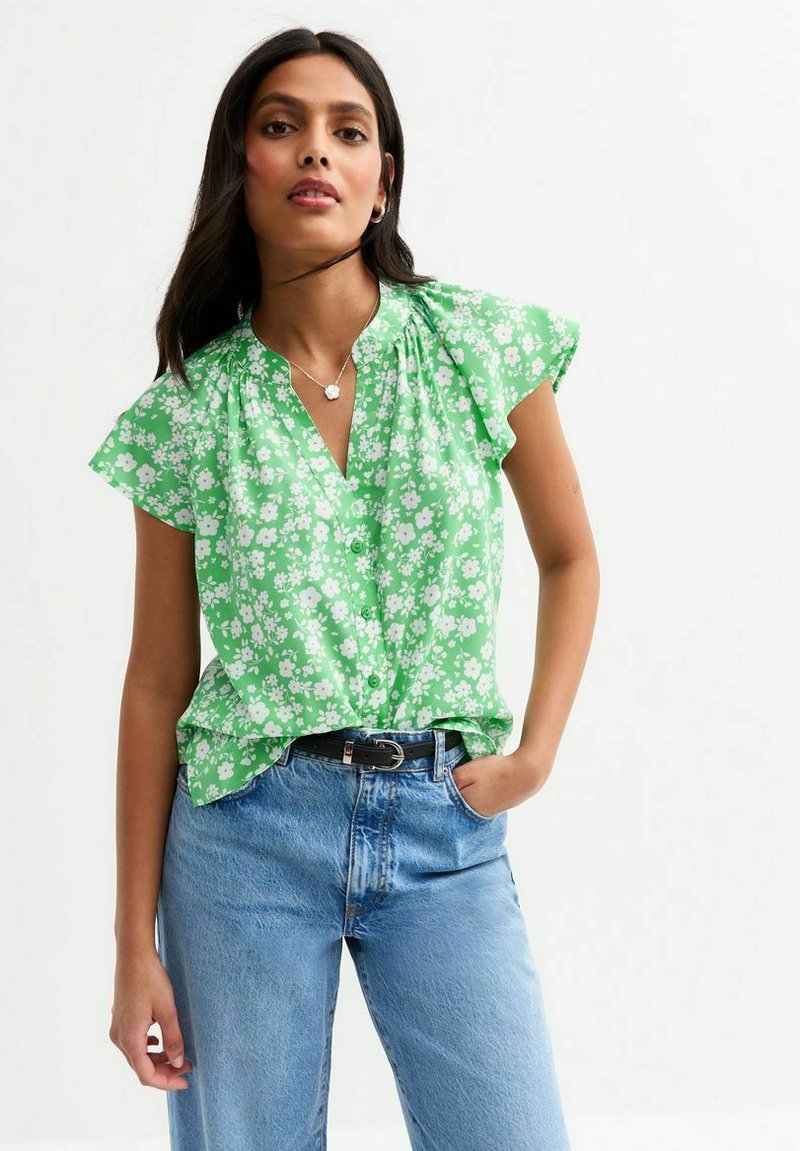 Blouse florale verte à manches courtes, avec un devant boutonné et un col froncé. Portée avec des jeans bleu clair retroussés et une ceinture noire.