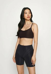 Ung kvinna med långt svart hår som bär en mörk sport-bh och högmidjade svarta cykelshorts mot en enkel bakgrund.