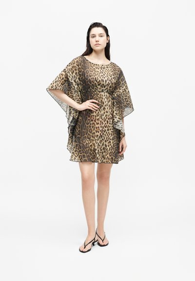 Νεαρή γυναίκα στέκεται με το χέρι στη μέση, φορώντας ένα διάφανο φόρεμα με λεοπάρ μοτίβο και μαύρα σανδάλια με τακούνι σε λευκό φόντο.