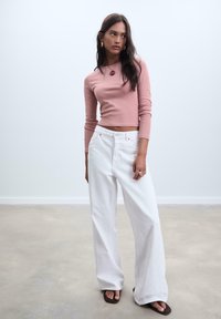 Růžový dlouhý rukáv crop top v kombinaci s volnými bílými širokými džíny. Model doplňuje kulatým přívěskem na náhrdelníku a prsteny.