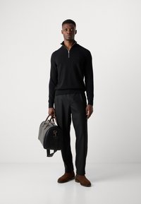 Zwarte sweater met rits, zwarte broek, bruine suède schoenen en een zwarte leren sporttas met bruine banden. Heeft een slim fit.