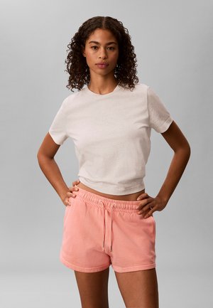 Kvinde med krøllet hår iført en hvid kortærmet top og lyserøde shorts med snøre, står med hænderne på hofterne foran en grå baggrund.