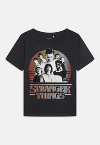 T-shirt graphique noir avec un imprimé circulaire représentant des personnages de "Stranger Things" et le titre en texte audacieux et coloré en bas.