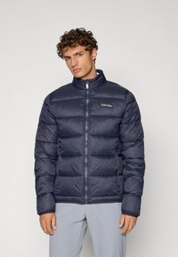 Marineblå dunjakke med kviltet design, høy krage, hel glidelås og sidelommer. Har Calvin Klein-logo på brystet. Lett materiale.