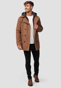 Brun dobbeltradet yderfrakke med sorte knapper og en grå hætteforing, parret med sorte slim-fit jeans og brune ankelstøvler.