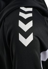 Tissu noir avec quatre flèches chevrons blanches pointant vers le bas en ligne verticale sur la zone de l'épaule d'un vêtement.