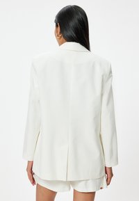 Witte blazer met lange mouwen, voorzien van een enkele achterkantopening, gestructureerd schouderdesign en een gladde stof, gestyled met lichte shorts.