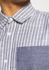 Skjorta med knappar i grå och vita vertikala ränder, en enfärgad grå ficka och en krage av lätt tyg.