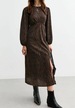 Femme portant une robe marron foncé à manches longues avec un imprimé animal noir, une fente latérale, des bottines noires et un collier avec un pendentif rond.