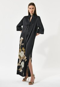 Kvinde i lang sort satin kjole med blomstermotiv og sidelæk, iført guld højhælede sandaler med blomsterdetaljer.