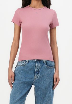 Frau, die ein enganliegendes, staubiges rosa T-Shirt mit kurzen Ärmeln und lockeren blauen Jeans mit Logodetails auf der Brust und der Tasche trägt.