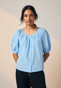 Hellblaue Bluse mit rundem Ausschnitt, kurzen Puffärmeln und Knopfverschluss, gefertigt aus strukturiertem Baumwollstoff mit dezenten, erhabenen Mustern.
