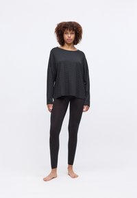 Mulher em pé, descalça, vestindo uma blusa oversized preta com padrão de mangas longas e leggings pretas ajustadas em frente a um fundo branco liso.