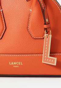 Sac à main en cuir orange avec une finition texturée, présentant des coutures blanches, un accent doré en forme de "L" et un logo de marque embossé "LANCEL PARIS."