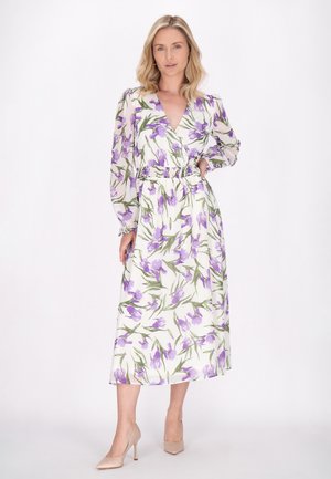 Witte jurk met paars bloemenpatroon, lange doorzichtige mouwen, een V-hals en een elastische taille. Gecombineerd met neutrale hoge hakken.