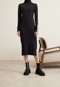 Svart långärmad polokragsklänning med en sidodelning, med kontrasterande gröna accenter på ärmarna. Bärs med klumpiga svarta ankelstövlar.