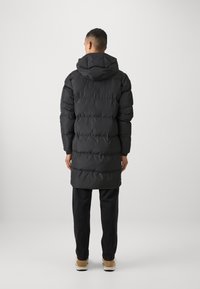 Svart dunjakke med hette, med horisontale quiltede seksjoner. Uformell passform, kombinert med mørke bukser og beige sko.