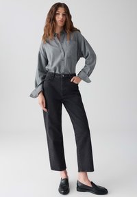 Donna dai capelli ondulati che indossa una camicia grigia a bottoni larga, jeans neri a vita alta e mocassini neri, in piedi con una mano in tasca.