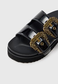 Crna sandala s patentnim remenom s dvije široke trake, zlatnim ukrasnim dizajnom i teksturiranom podlogom za stopala radi prianjanja. Potplat na platformu.
