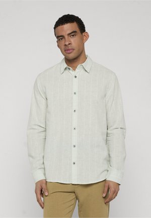 Mężczyzna ubrany w jasnozieloną, długorękawową koszulę na guziki i musztardowe spodnie, stojący na tle jednolitego szarego tła.