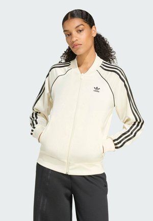 Cream zip-up jacket made from smooth fabric, featuring black stripes on sleeves and a small black logo on the chest. Pairing with dark pants.