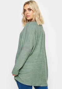 Långärmad grön tröja med texturerad yta, prydd med nitar vid axlarna och med en avslappnad passform.