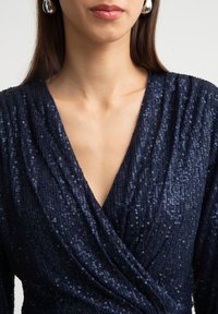 Jūras zilas, sequined tops ar V-veida kakla izgriezumu un savāktu audumu. Tuvplāns uz krūšu daļu, izceļot spīdumu un tekstūras detaļas.